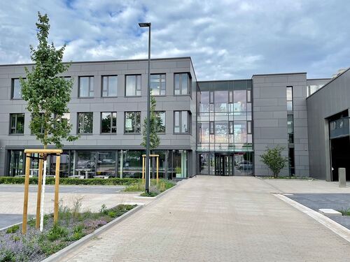 Blick von der Zufahrt aus auf die Eingangsfassade des Gebäudes mit Parkplätzen, Bäumen und Himmel