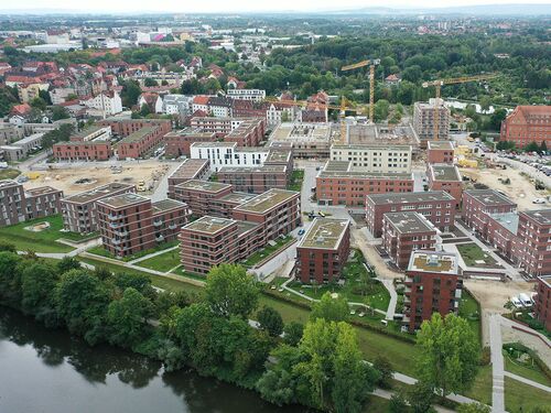 Blick von oben auf die vielen roten Baukörper, eingebettetet in Grünanlagen entlang des Flusses mit Bäumen