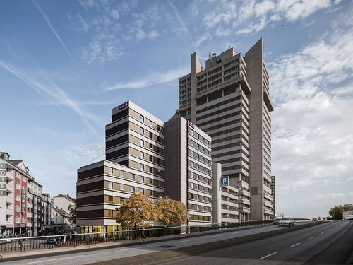 Blick auf den großen, grauen Gebäudekomplex mit Hochhaus im hinteren Bereich und Schnellstraße im Vordergrund