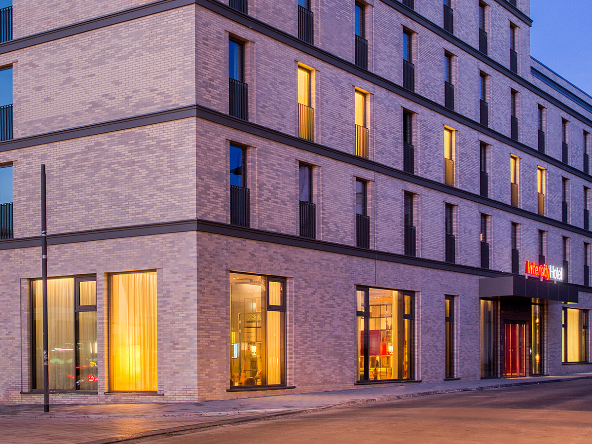 Abendlichtansicht auf dei Ecke des Gebäudes in rot schimmerndem Licht und mit warmer Beleuchtung hinter den Fenstern