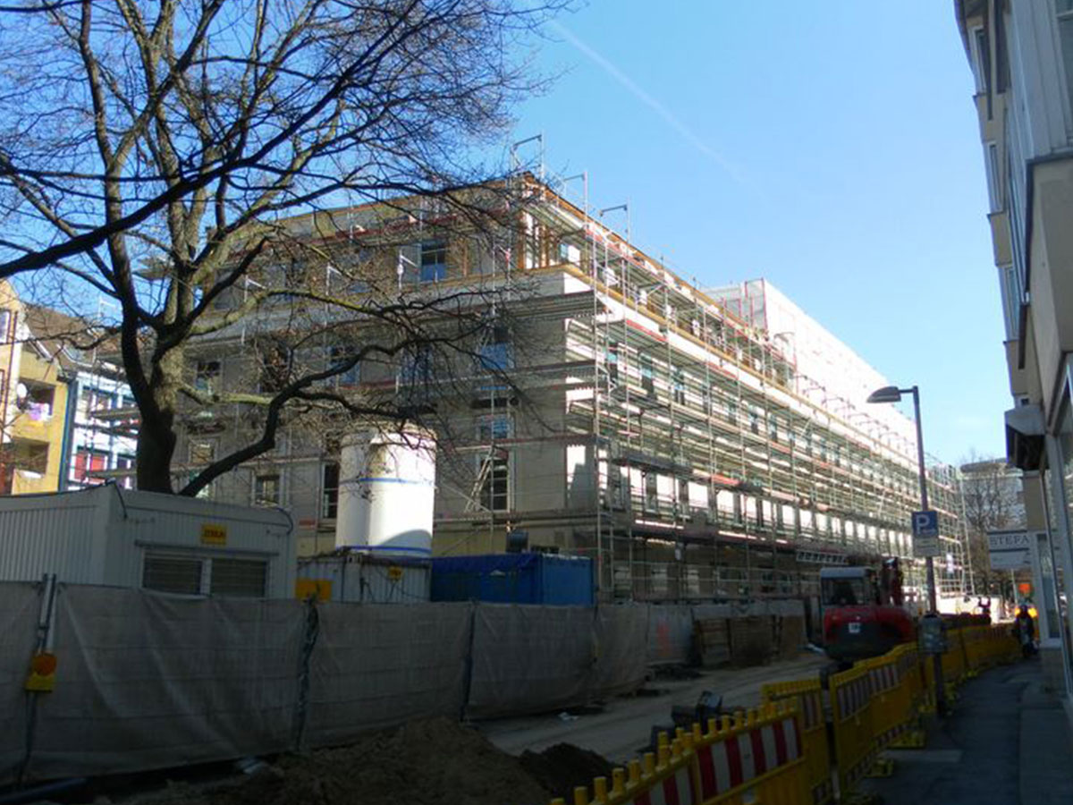 Baustellenbild des eingerüsteten Rohbaus mit laubfreiem Baum und vielen Bauzaunabsperrungen und Containern