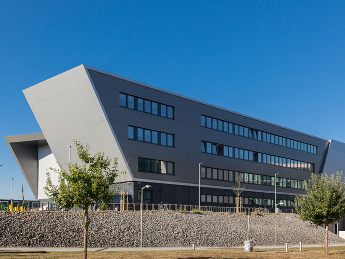 Blick auf das trapezförmige, graue und futuristische Gebäude mit Kieswall, Bäumen und Begrünung im Vordergrund